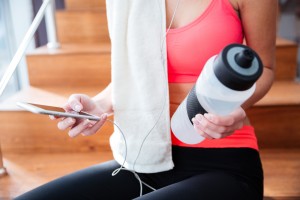 Closeup of young sportswoman sitting and holding cell phone and bottle of water in gym